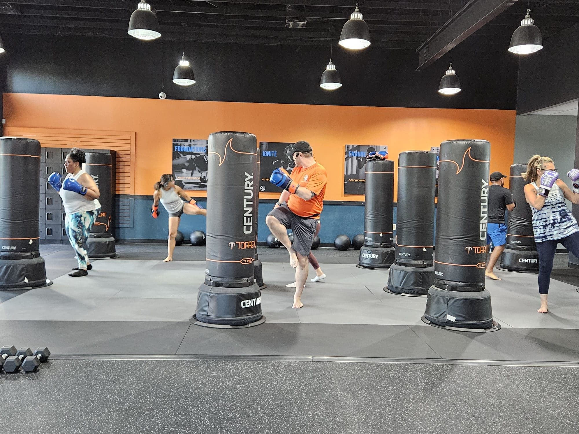 A Vibes class mid-round, members kicking heavy bags while a coach in orange demonstrates
